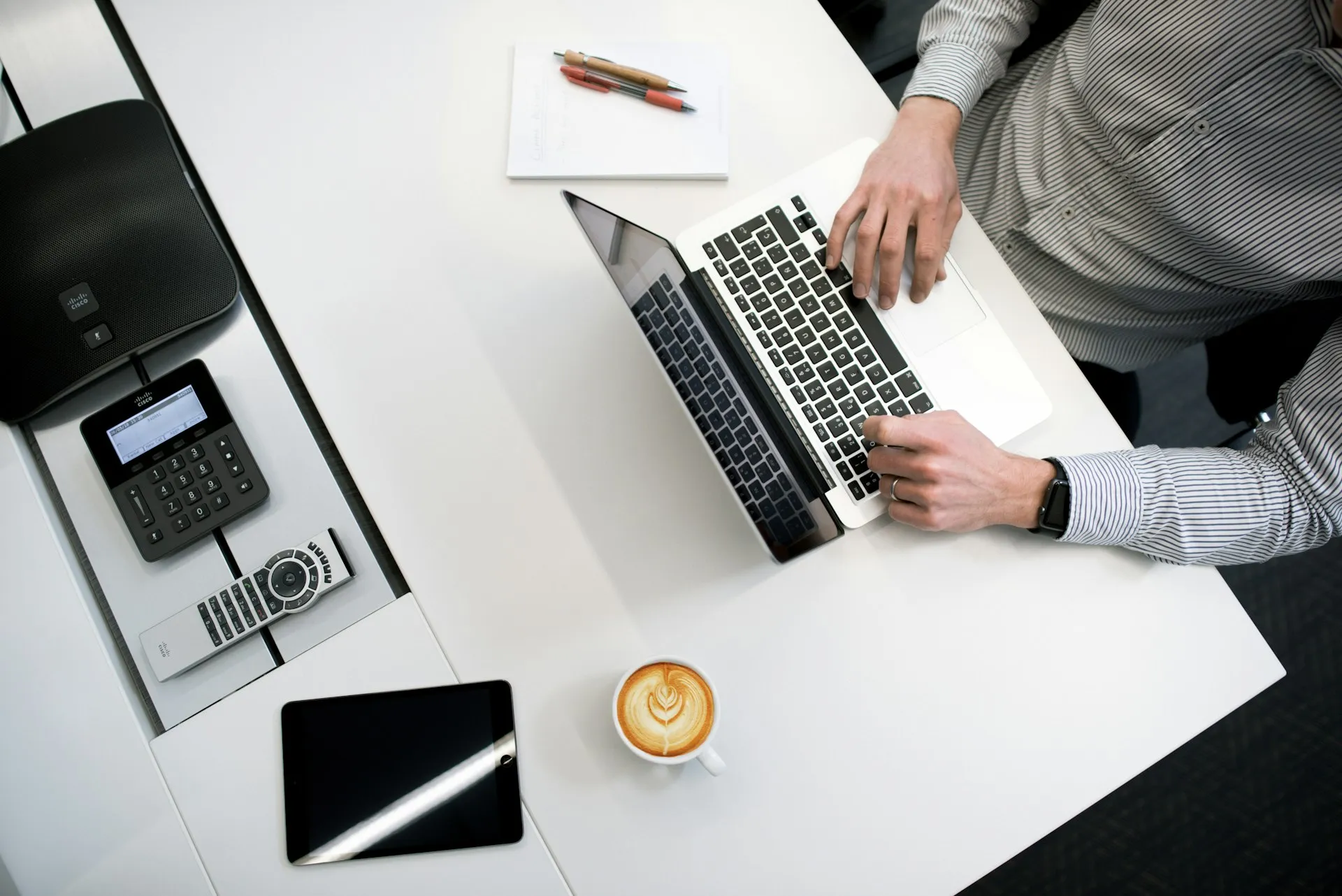 Person working on a laptop Overhead view of a person working on a laptop at a clean white desk, with a notebook, tablet, latte, and office phone nearby.