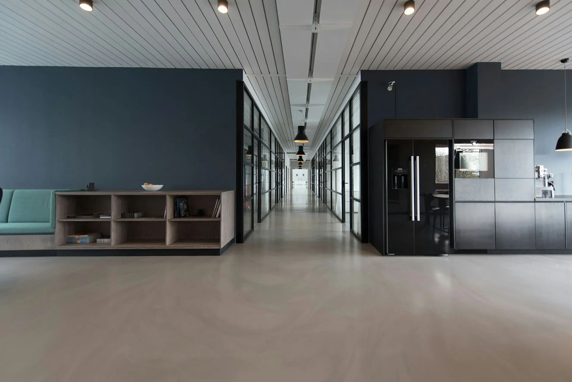 Modern office interior Modern office interior with a long hallway flanked by glass-walled rooms, a lounge area on the left, and a sleek black kitchen area with pendant lighting on the right.